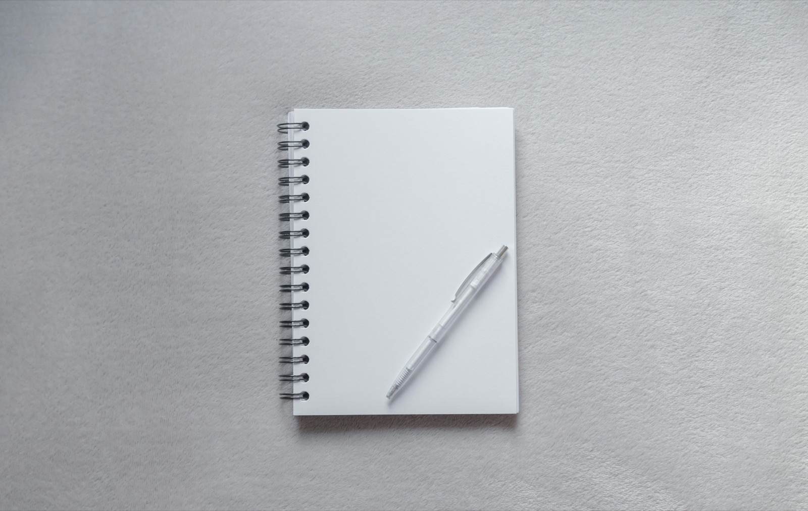 A blank white spiral notebook and a translucent pen on a grey surface; illustrative stock photograph.