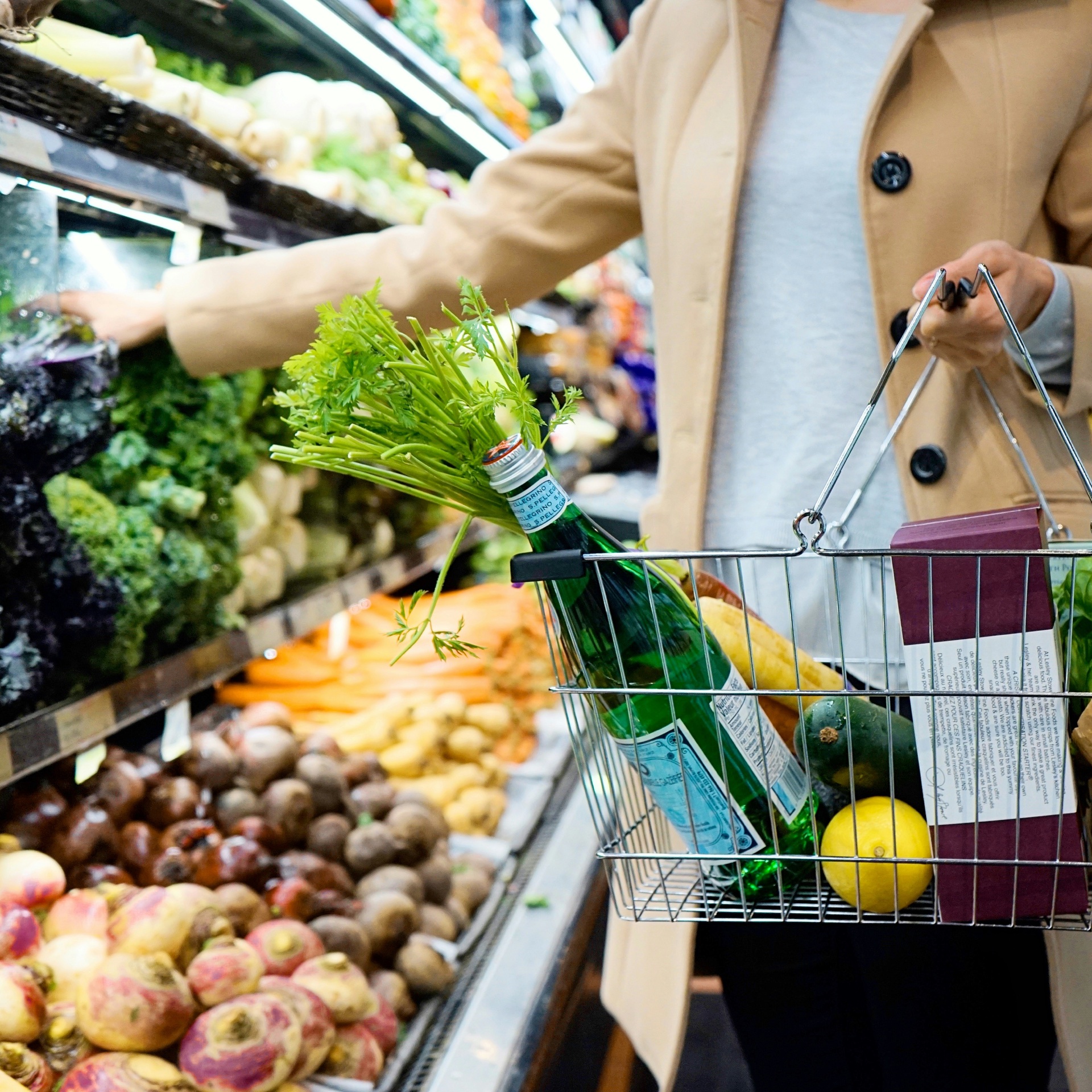 A shopper carrying a wire basket of groceries beside a supermarket produce display.